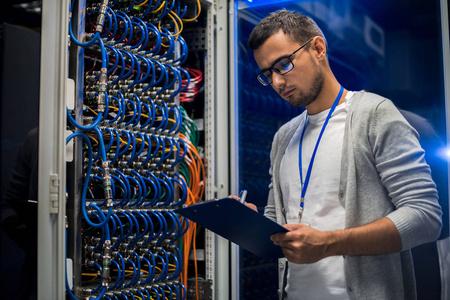 年轻的网络工程师站在服务器机柜旁边,同时在数据中心与超级计算机